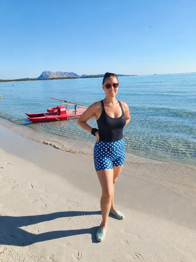 Tessa Brooks in swimwear on a sandy beach, with a red rescue boat in the shallow water behind her